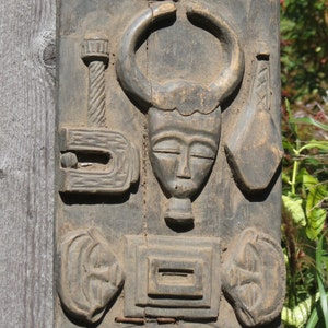 May include: A dark wood carving of a bull's head with a human face. The carving is detailed and intricate, with a variety of shapes and patterns. The carving is mounted on a wooden board.