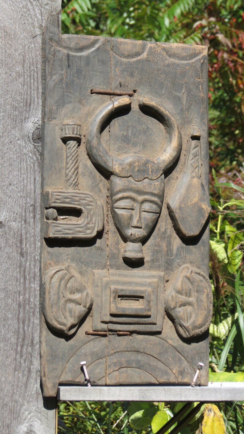 May include: A dark wood carving of a bull's head with a human face. The carving is detailed and intricate, with a variety of shapes and patterns. The carving is mounted on a wooden board.