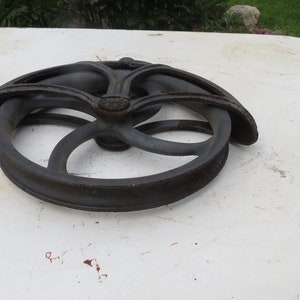 May include: A black cast iron well pulley with a decorative design. The pulley has a grooved rim and a central hub with a decorative design.