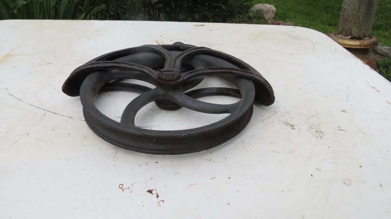 May include: A black cast iron well pulley with a decorative design. The pulley has a grooved rim and a central hub with a decorative design.