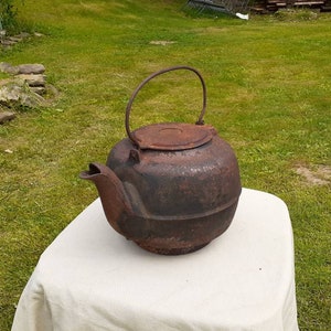 May include: A vintage cast iron kettle with a dark brown patina. The kettle has a lid and a handle. It is sitting on a white tablecloth.