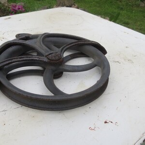 May include: A cast iron pulley wheel with a decorative design. The wheel is dark gray and has a smooth, worn surface.