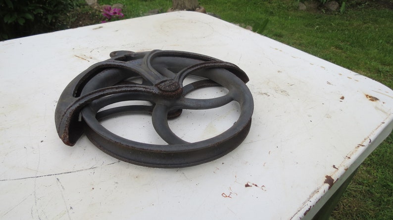May include: A cast iron pulley wheel with a decorative design. The wheel is dark gray and has a smooth, worn surface.