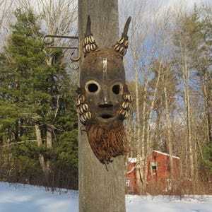 May include: A wooden mask with a dark brown finish and a beard made of woven fibers. The mask has a human-like face with large eyes and a wide mouth. The mask is decorated with small, white shells around the eyes and forehead.