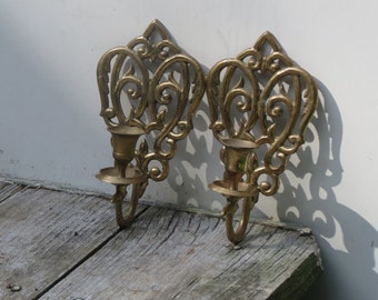 May include: Two ornate gold metal candle holders with a scrolled design. The candle holders are mounted on a white wall.