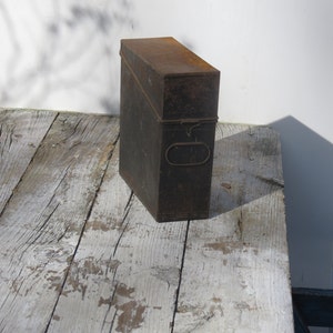 May include: A vintage metal box with a rusty brown finish and a metal latch. The box is sitting on a weathered wooden surface.