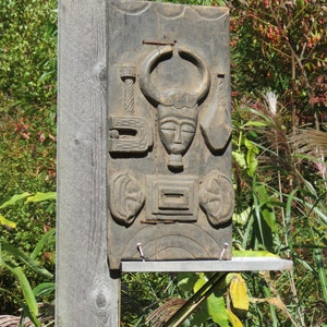 May include: A dark wood carving of a face with horns, set against a wooden background. The carving is mounted on a wooden post.