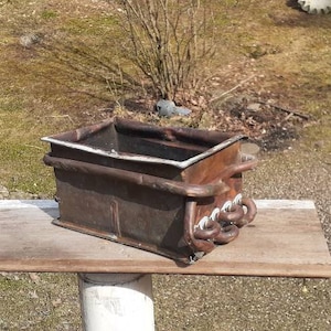 May include: A rusty metal planter with a rectangular shape and a white interior. The planter has a decorative metal design on the front and is sitting on a wooden surface.