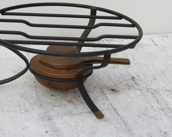 May include: A black metal stand with a round top and three legs. The stand has a copper pot with a lid on top. The stand is on a white surface.