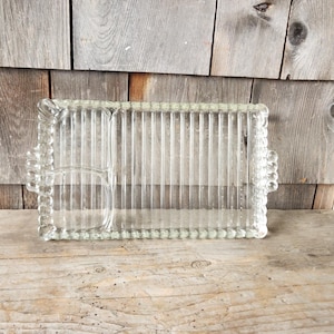 May include: A clear glass serving tray with a rectangular shape and a small, round compartment. The tray has a ribbed design and beaded edges. It sits on a wooden surface.
