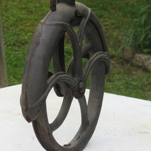 May include: A vintage cast iron pulley wheel with a decorative design. The wheel is dark gray with rust spots and has a metal loop at the top.