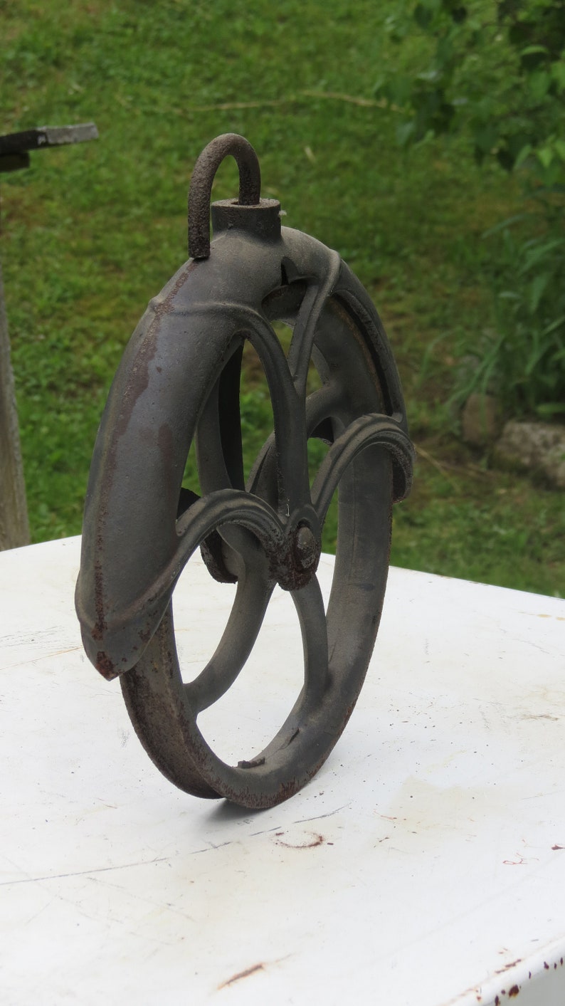 May include: A vintage cast iron pulley wheel with a decorative design. The wheel is dark gray with rust spots and has a metal loop at the top.