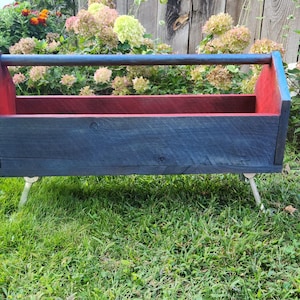 May include: A wooden toolbox painted blue and red with a wooden handle. The toolbox is sitting on three metal legs.