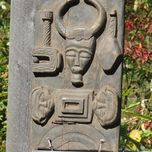 May include: A dark wood carving of a face with horns, surrounded by other carved shapes. The carving is mounted on a wooden board.
