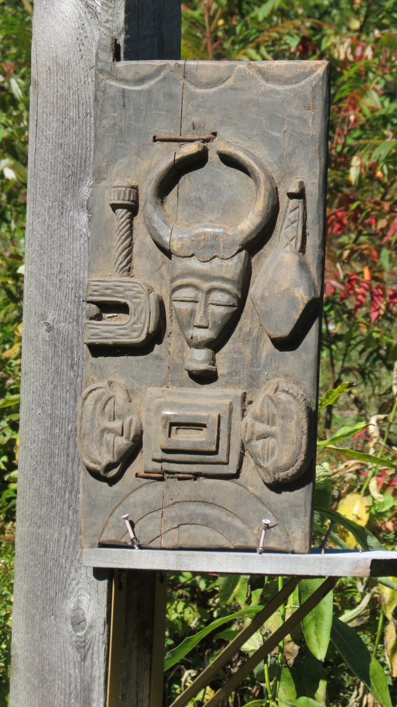 May include: A dark wood carving of a face with horns, surrounded by other carved shapes. The carving is mounted on a wooden board.