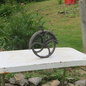 May include: A black metal pulley wheel with a decorative design sits on a white table. The pulley is hanging from a hook and has a diameter of approximately 12 inches.