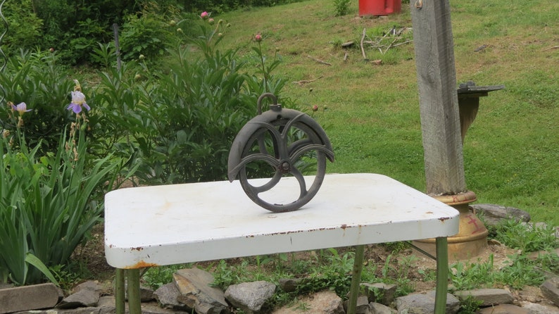 May include: A black metal pulley wheel with a decorative design sits on a white table. The pulley is hanging from a hook and has a diameter of approximately 12 inches.