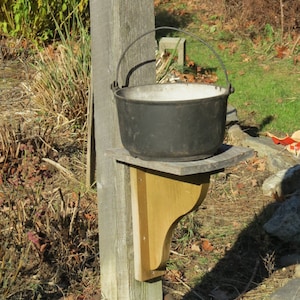May include: A rustic outdoor setup featuring a black cast iron pot with a handle, resting on a wooden shelf attached to a weathered wooden post. A golden metal base with red lettering is at the post's base.