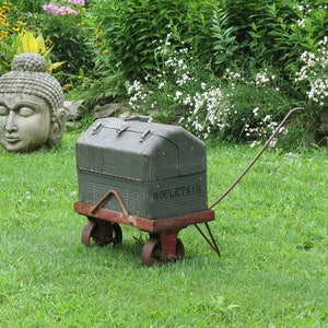 May include: A green metal toolbox on a red wooden cart with black wheels. The cart has a long metal handle. The toolbox has the word "KNOBBY" printed on the side. The cart is sitting on a green grassy lawn in front of a stone wall and a garden.