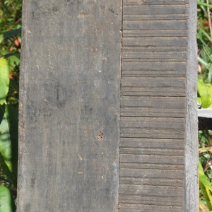 May include: A weathered wooden panel with a grooved section on the right side. The panel has several rusty nails and a metal loop at the top.
