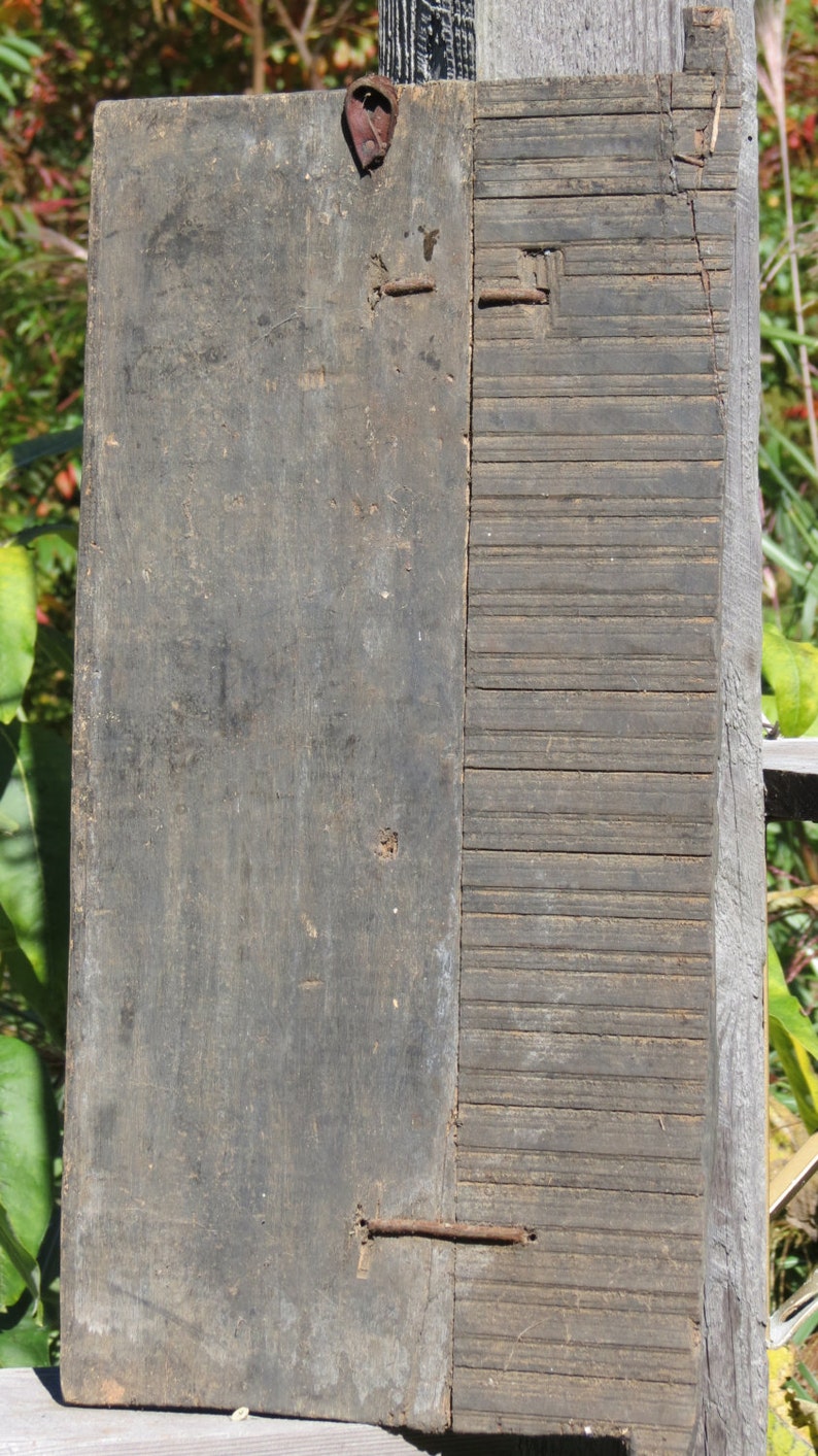 May include: A weathered wooden panel with a grooved section on the right side. The panel has several rusty nails and a metal loop at the top.