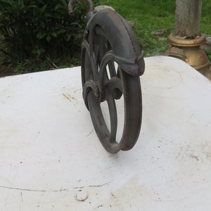 May include: A black cast iron pulley wheel with a hook. The wheel has a decorative design and is sitting on a white surface.