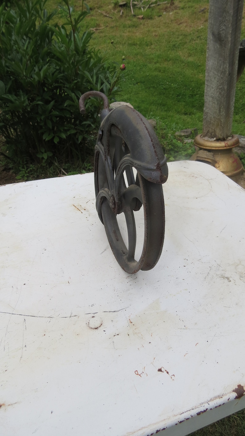 May include: A black cast iron pulley wheel with a hook. The wheel has a decorative design and is sitting on a white surface.