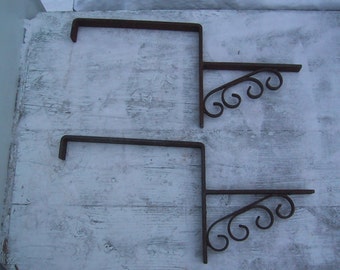 May include: Two rusty metal brackets with decorative scrollwork on the ends. The brackets are designed to be mounted to a wall or other surface.