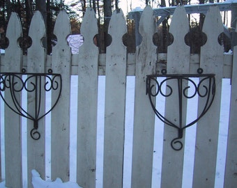 May include: Two black metal plant hangers with a decorative scroll design are attached to a white picket fence. The fence is made of vertical wooden slats with a rounded top.