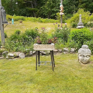 May include: A stone Buddha head sculpture sits on the grass in front of a wooden table with a planter box filled with green plants. The table is in a garden setting with a stone wall and green grass.