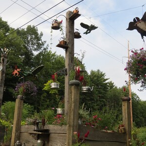 May include: A wooden garden structure with multiple wooden posts and a variety of hanging teapots and other metal objects. The structure is decorated with flowers and plants.