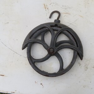 May include: A black cast iron pulley with a decorative design and a hook. The pulley has a diameter of approximately 8 inches.