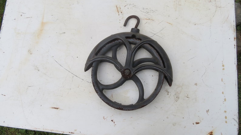 May include: A black cast iron pulley with a decorative design and a hook. The pulley has a diameter of approximately 8 inches.