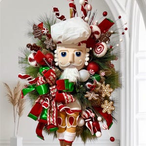 May include: A festive Christmas decoration featuring a nutcracker chef design. The decoration is adorned with red, green, and white ribbons, gingerbread cookies, candy canes, and ornaments. The chef wears a white hat and has a detailed face.