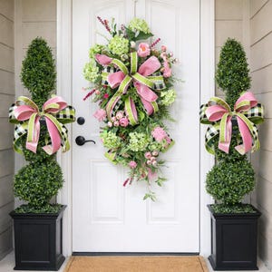 May include: A white front door is decorated with a floral wreath and two topiary trees. The wreath and topiaries are adorned with pink, green, and black and white checkered bows. The topiaries are in black square planters.