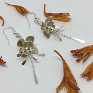 Può includere: Un paio di orecchini d'argento a forma di fiori delicati con steli pendenti. Gli orecchini sono esposti su una superficie bianca con petali di fiori arancioni secchi.
