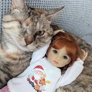 Puede incluir: Una muñeca de pelo rojo con una camisa blanca de temática navideña con un diseño de Papá Noel y un reno, tumbada en un sofá a rayas grises y blancas con un gato atigrado gris y marrón.