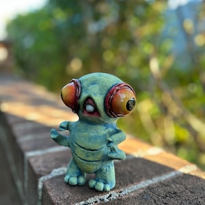 May include: A small, green figurine of an alien-like creature with large, orange and red eyes. The figure has small arms and feet, and is standing on a brick surface. The background is blurred, with green foliage.