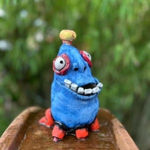 May include: A small, hand-sculpted blue creature figurine with a textured surface, featuring large white eyes with red rims, a wide mouth with visible white teeth, a yellow knob on top of its head, and four short, stubby red legs at the base.