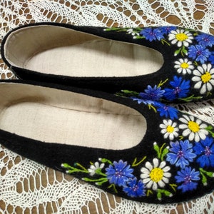 Puede incluir: Par de zapatillas de fieltro negro con flores blancas y azules bordadas. Las zapatillas tienen un forro blanco.