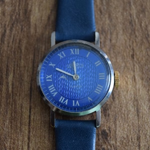 May include: A blue wristwatch with Roman numeral hour markers and a textured blue face. The watch has a silver case and a blue leather band.