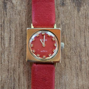 May include: A gold-toned square wristwatch with a red face and a red leather band. The watch face has the word "antibio" printed on it.