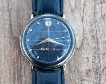 Puede incluir: Un reloj de pulsera negro y dorado con una correa de cuero negro. La esfera del reloj tiene un fondo negro con marcas blancas y la palabra "PAKETA" en letras doradas.