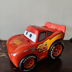 May include: A red toy race car featuring the number 95 on the side, Rust-eze branding on the hood and rear spoiler, and black tires with Lightyear lettering. The car has white eyes on the windshield and orange wheels with a stylized design.