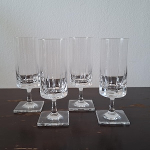 May include: Four clear, stemmed glasses with a rectangular base. The glasses have a tall, cylindrical shape with vertical etched lines. The glasses are arranged on a dark brown surface against a white wall.