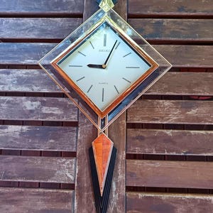 May include: A vintage Seiko quartz wall clock with a geometric design. The clock has a square face with black hands and hour markers, in a gold-toned frame. The clock has a decorative, elongated pendant with orange, black, and gold accents. The clock is mounted on a wooden surface.