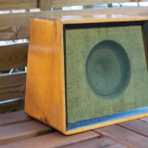 K&ouml;nnte beinhalten: Ein Vintage-Lautsprecher aus Holz mit hellbraunem Finish. Der Lautsprecher hat eine gr&uuml;ne Netzfront und eine kreisf&ouml;rmige Lautsprecherkomponente, die hinter dem Netz sichtbar ist. Der Lautsprecher ist rechteckig mit trapezf&ouml;rmiger Form.