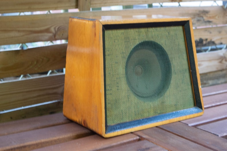 K&ouml;nnte beinhalten: Ein Vintage-Lautsprecher aus Holz mit hellbraunem Finish. Der Lautsprecher hat eine gr&uuml;ne Netzfront und eine kreisf&ouml;rmige Lautsprecherkomponente, die hinter dem Netz sichtbar ist. Der Lautsprecher ist rechteckig mit trapezf&ouml;rmiger Form.