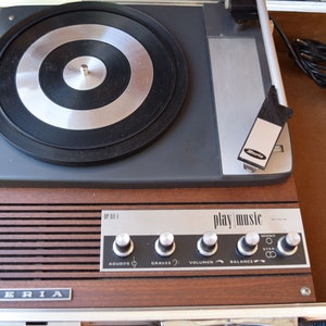 May include: A vintage brown and silver portable record player with a black turntable and a silver tone arm. The player has a woodgrain finish and a silver metal nameplate that reads "IBERIA". The player has a control panel with knobs labeled "Agudos", "Graves", "Volumen", "Balance", and "Mono Stereo". The player also has a label that reads "Play Music".