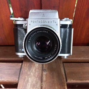 May include: A silver Pentacon Six TL camera with a black leather grip and a Carl Zeiss Jena DDR lens. The camera is set against a wooden background.